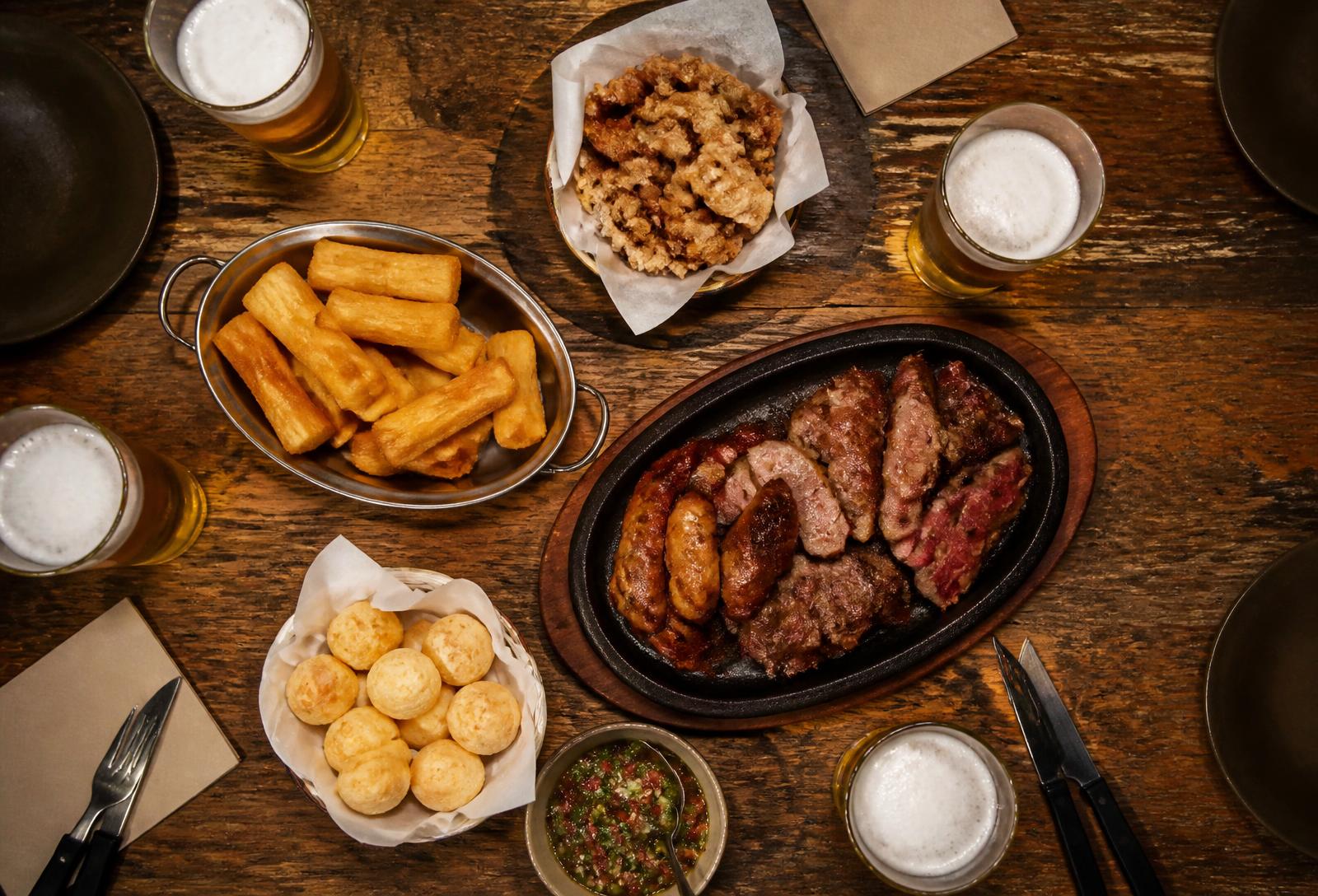 Mesa de madeira vista de cima com chopp, porções, mandioca frita, pão de queijo e talheres